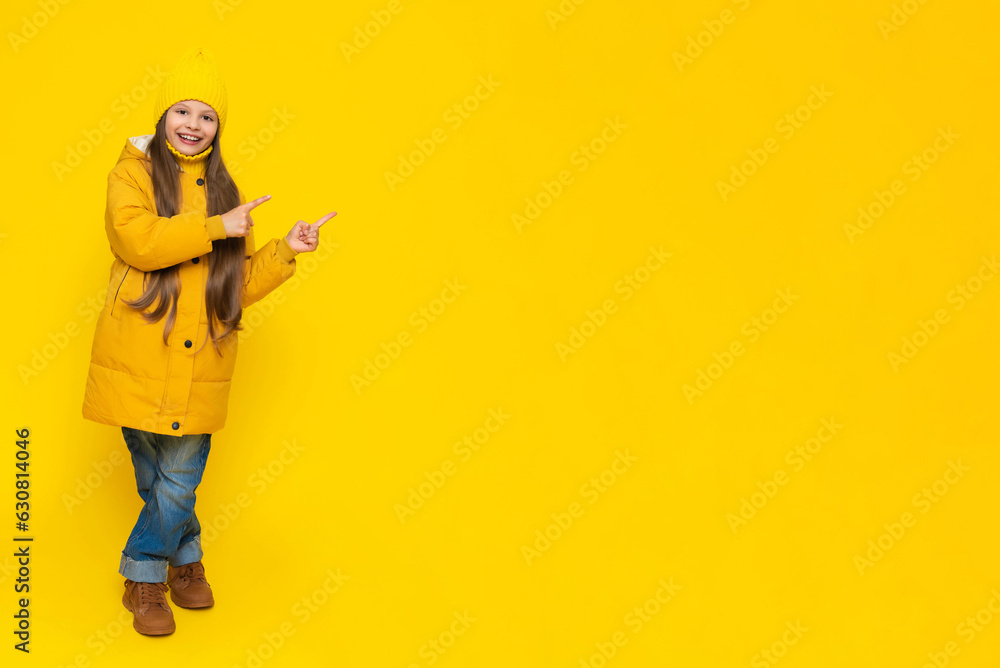 A full-length little girl in a jacket and hat points to the announcement of your advertisement. The child rejoices in autumn. Warm clothes for children. Yellow isolated background. Copy space. A full-length little girl in a jacket and hat points to the announcement of your advertisement. The child rejoices in autumn. Warm clothes for children. Yellow isolated background. Copy space.