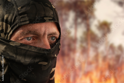 Military man in a camouflage mask against the backdrop of a burning forest. The concept of military action. Man at war. Fire after explosions. Forest fire. Arsonist. Closeup of the face.