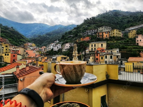 A cup of coffee against the background of the ancient and medieval city of Riomaggiore in the Cinque Terre National Park. Relaxing view of cityscape with colorful houses in gloomy weather