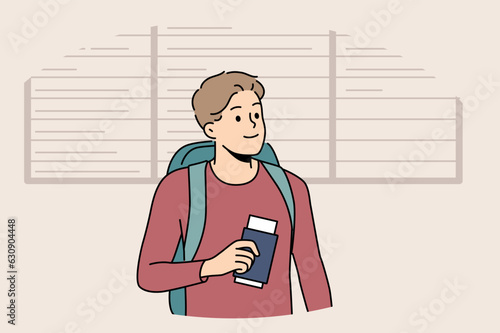 Man tourist stands at airport near board of upcoming flights and holds passport and plane ticket. Guy passenger of airport with backpack behind back is going on trip and waiting for flight