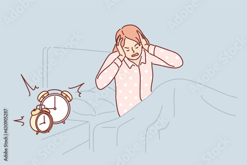 Woman wakes up with difficulty early in morning sitting in bed near bedside table with alarm clock