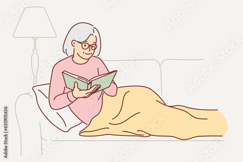 Grandmother is reading book sitting on couch, covering legs with blanket and enjoying exciting plot