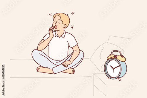 Boy yawns and wants to sleep sitting on bed near alarm clock and feels morning sleepiness