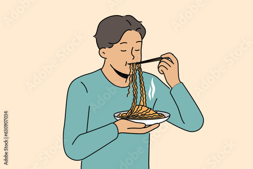 Asian man eats ramen with chinese sticks, enjoying taste of traditional national meal from korean cuisine restaurant. Guy stands and enjoys eating ramen or noodles to get enough during work break