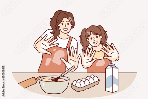 Mom and daughter are preparing breakfast or pastries and with smile show palms stained with flour. Woman brings up little girl teaching how to cook pastries from dough and enjoys raising child