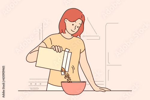 Woman is eating breakfast with cornflakes and holding box of muesli standing near kitchen table