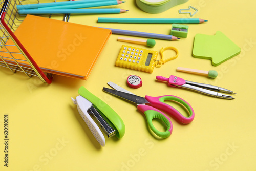 Shopping basket with different school stationery on yellow background, closeup