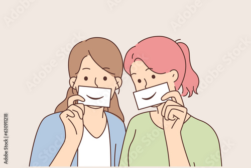 Two women with fake smiles hold papers with drawn emotions and pretend to be happy