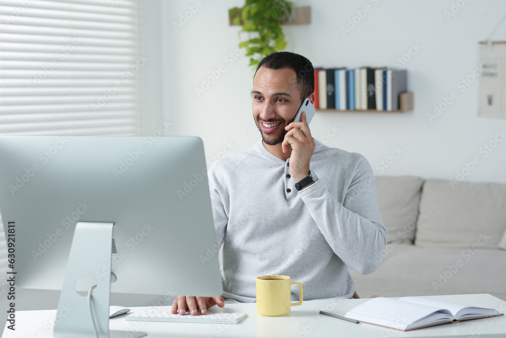 Young man talking on smartphone while working with computer at desk in room. Home office