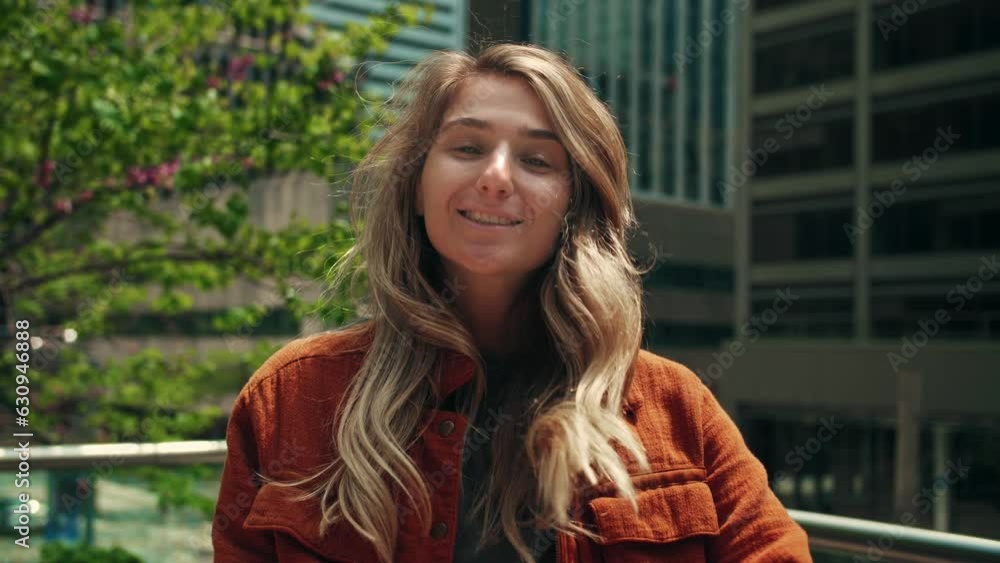 Blonde woman smiling at camera on city background.Young lady fixes hair standing against a beautiful city and trees.Female wearing orange jacket looking at camera in the great mood. Blonde woman smiling at camera on city background.Young lady fixes hair standing against a beautiful city and trees.Female wearing orange jacket looking at camera in the great mood.