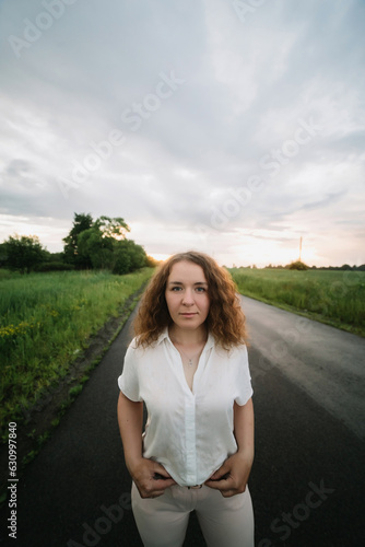 Young woman stands on the road. Young woman in hat with curly hair. Sunset or sunrise, bright evening light