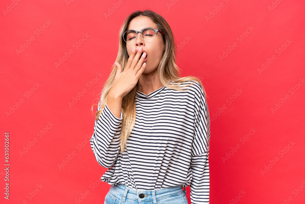 Young Uruguayan woman isolated on red background yawning and covering wide open mouth with hand Young Uruguayan woman isolated on red background yawning and covering wide open mouth with hand