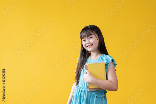 An adorable Asian girl holding her books while standing against an isolated yellow background.