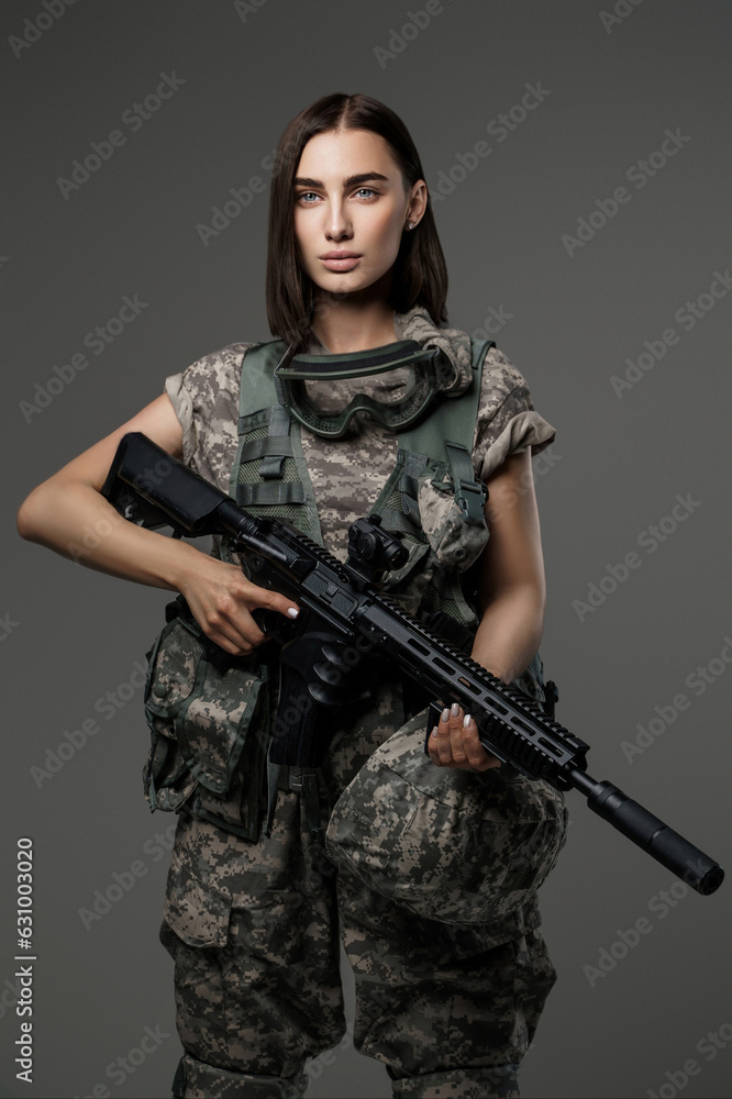 Beautiful female soldier with stunning blue eyes stands in her military uniform holding an automatic rifle for a studio portrait taken against a grey background. Beautiful female soldier with stunning blue eyes stands in her military uniform holding an automatic rifle for a studio portrait taken against a grey background.