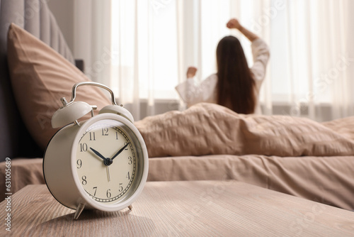 It's lazy morning o'clock. Alarm clock on bedside table and woman stretching in room, selective focus