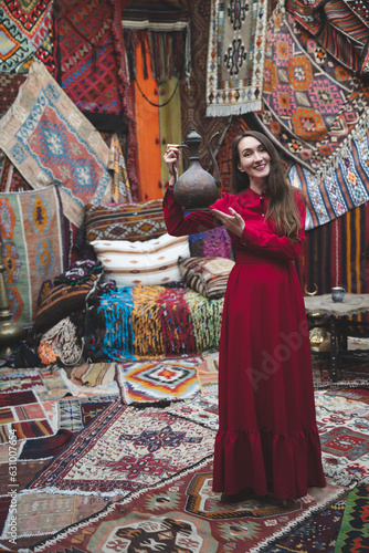 beautiful girl in a red dress with a teapot in a traditional turkish interior with many carpets on the wall
