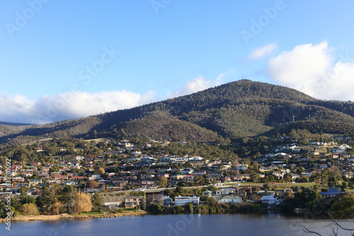 Residential houses nestled at the bottom of a large mountain with a cloudy blue sky on the Derwent River in Tasmania 