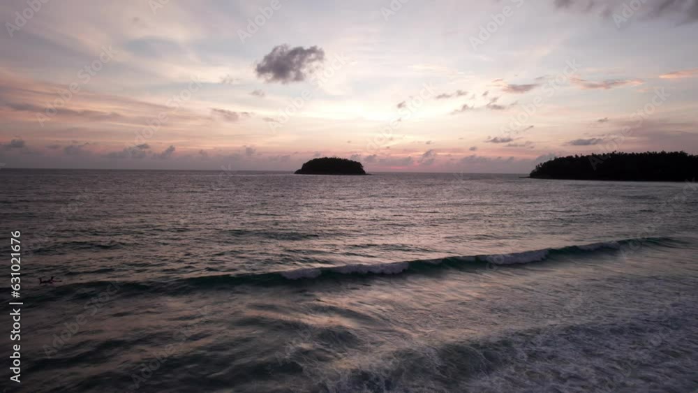 A lonely island stands in the ocean at sunset. The view from the drone. Small waves flow around it creating ripples on the surface of ocean. The water is like a black mirror. Pinkish dark clouds are A lonely island stands in the ocean at sunset. The view from the drone. Small waves flow around it creating ripples on the surface of ocean. The water is like a black mirror. Pinkish dark clouds are