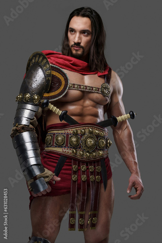 Powerful gladiator with a stylish beard wears lightweight intricate armor and poses gripping a feathered helmet against a grey background