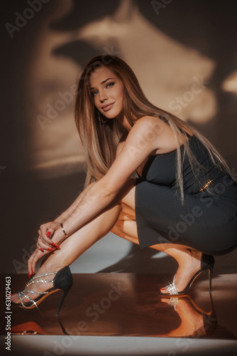 A stunning woman in a black evening gown gracefully poses in a squatting position on a golden reflective floor, beautifully accentuating the light in the background