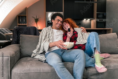 Interracial couple sitting on couch, snuggled in blankets, eating popcorn and scrolling through movies on television while talking