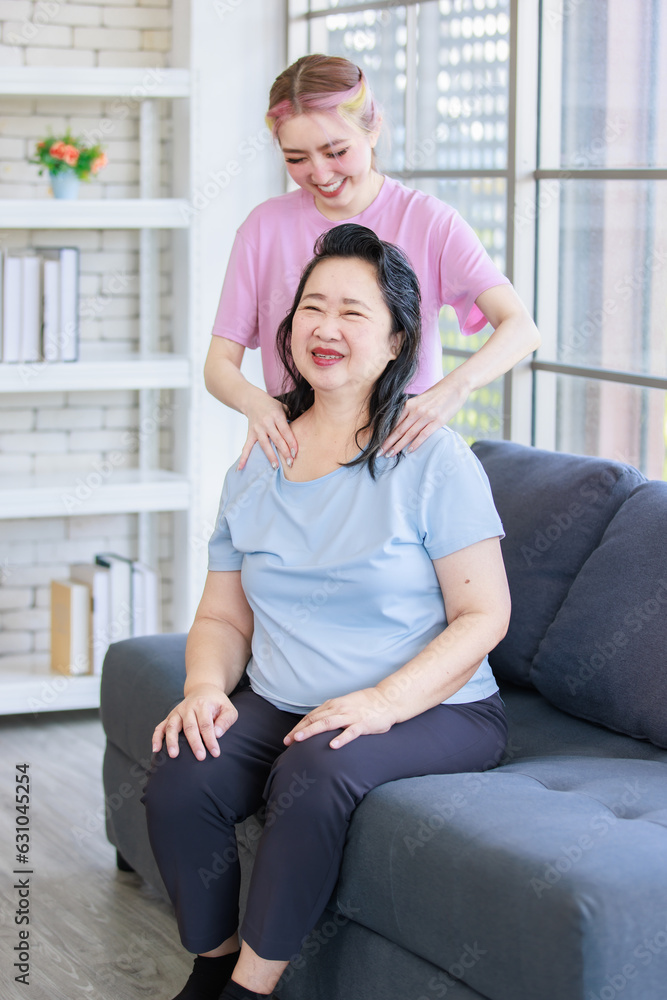 Obraz premium Asian young beautiful daughter standing behind massaging neck of old senior pensioner retirement cheerful happy healthy fit mother in casual sportswear sitting on sofa couch in living room at home