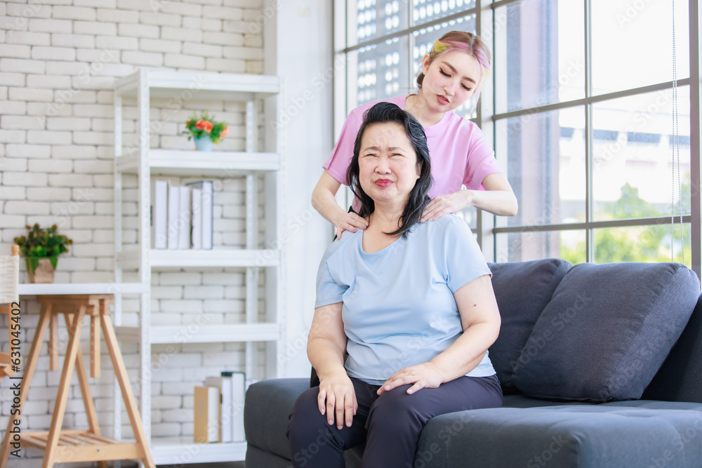 Asian young beautiful daughter standing behind massaging neck of old senior pensioner having backache from stretching in casual sportswear sitting on sofa couch in living room at home. Asian young beautiful daughter standing behind massaging neck of old senior pensioner having backache from stretching in casual sportswear sitting on sofa couch in living room at home.