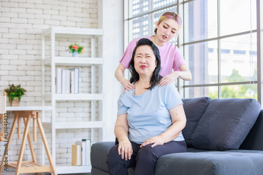 Obraz premium Asian young beautiful daughter standing behind massaging neck of old senior pensioner having backache from stretching in casual sportswear sitting on sofa couch in living room at home.