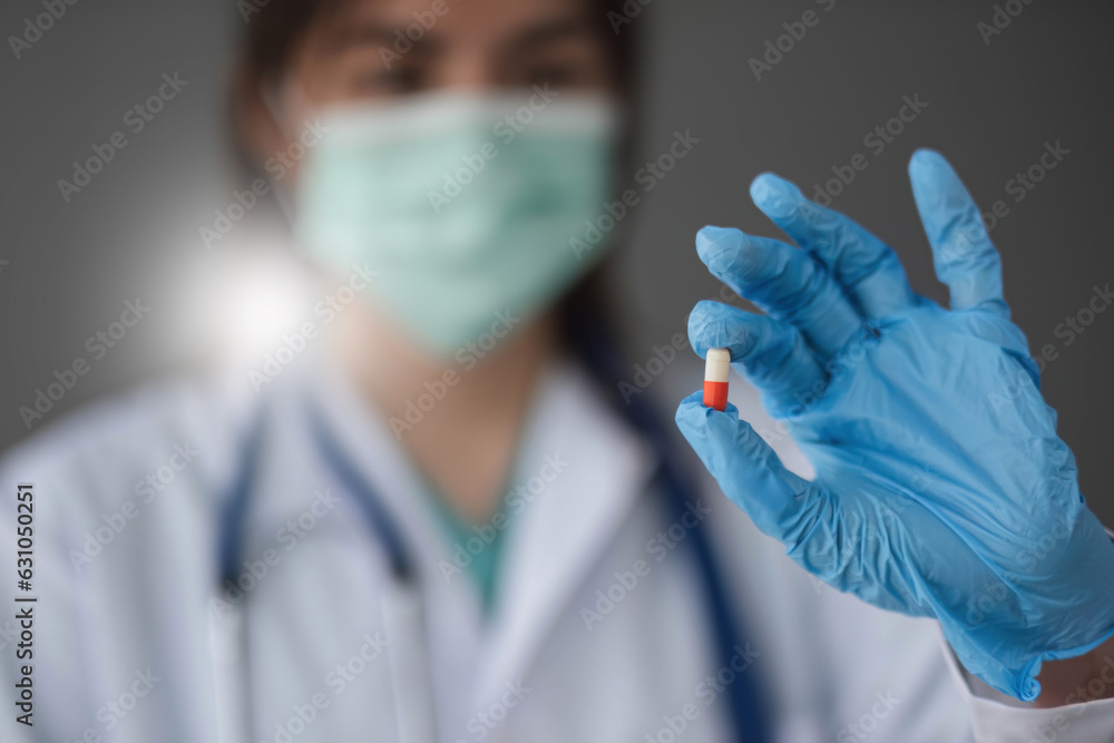 Close up of masked pharmacy doctor holding red transparent pill with finger in glove in pharmaceutical research laboratory Close up of masked pharmacy doctor holding red transparent pill with finger in glove in pharmaceutical research laboratory