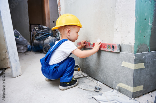 Little boy construction worker measuring wall surface with spirit level tool. Kid in work overalls using bubble level instrument while preparing wall for repair works at home during renovation.