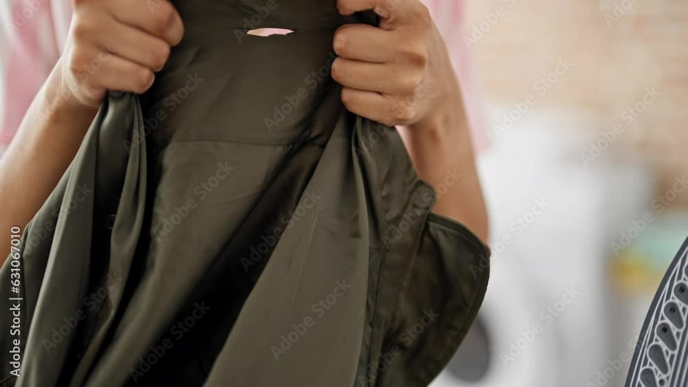 Young beautiful hispanic woman holding shirt with hole ironing at laundry room Young beautiful hispanic woman holding shirt with hole ironing at laundry room