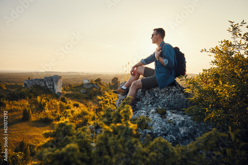 Travel and hiking concept. Adult man with backpack sitting on the edge of rock enjoying landscape during sunset