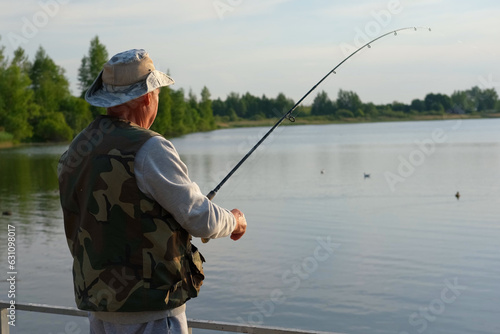 Old man fishing. Senior gray haired fisherman throws a spinning from shoreside at sunset, twisting a coil. Positive elderly male angling at lake, rotating reel rod. Retired fisher enjoying his weekend