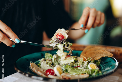 Pieces of caesar salad on fork supported by knife in female hands over plate with food on blurred background