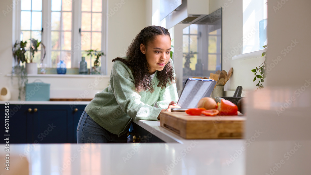 Teenage Girl At Home In Kitchen With Ingredients Looking At Recipe On Digital Tablet Teenage Girl At Home In Kitchen With Ingredients Looking At Recipe On Digital Tablet