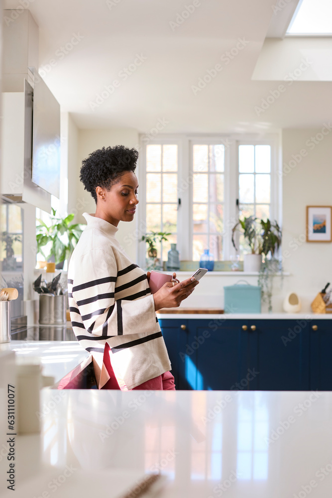 Mature Woman At Home In Kitchen Drinking Coffee And Checking Social Media On Mobile Phone Mature Woman At Home In Kitchen Drinking Coffee And Checking Social Media On Mobile Phone