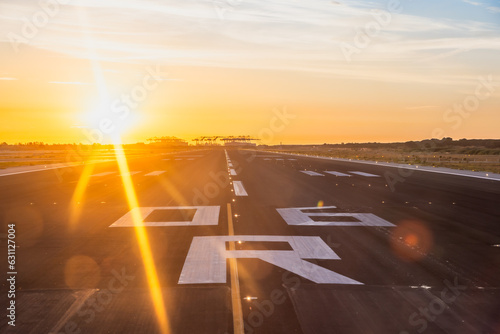 Cockpit view of tarmac runway shortly before departure. Bright yellow sunrise just above the horizon creating sun flares. Cranes of harbour visible in the distance. 