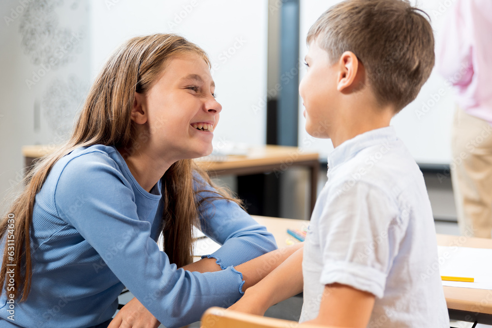 Cheerful schoolchildren at their desks sit at recess chatting and laughing. The schoolgirl smiles and looks at her classmate, he tells a funny story. Chatter and conversation during the lesson