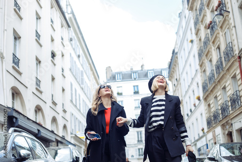 Photography Portrait of two young good-looking women wearing black trench coats, walking along street between cars, holding hands