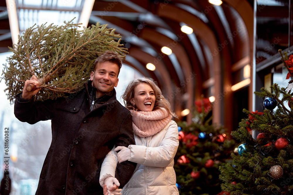 Beautiful happy young couple in love walking around Christmas market. Caucasian man and woman just bought Christmas tree and carry it home to decorate. Anticipation of the New Year Eve in snowy city.