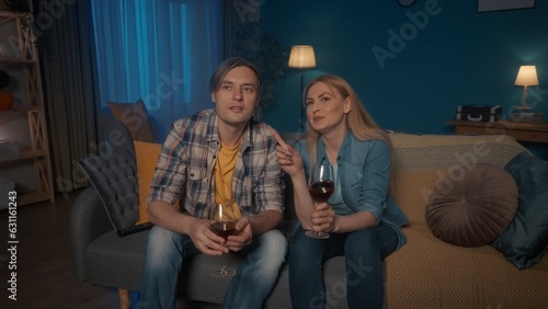 A young couple is sitting on a sofa with glasses of red wine and watching TV. A man and a woman are watching a movie with interest, a touching video. Weekend leisure concept.