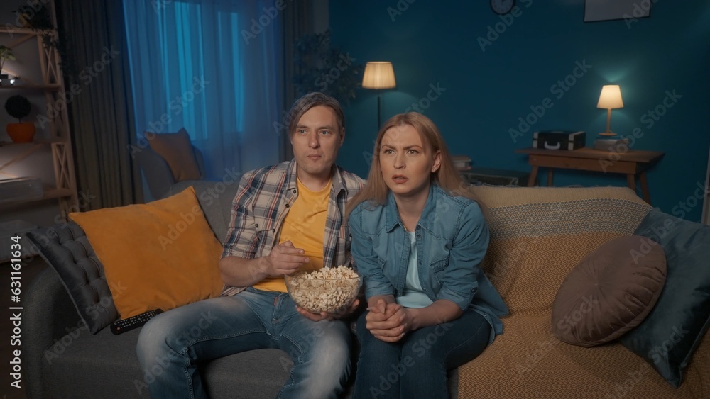 A man and a woman are watching a horror movie while sitting on the couch with a plate of popcorn. A married couple is closely following the plot with a surprised expression on their faces. A man and a woman are watching a horror movie while sitting on the couch with a plate of popcorn. A married couple is closely following the plot with a surprised expression on their faces.