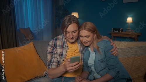 A young married couple sits at home on the couch and looks at the smartphone in the man's hand. A man and a woman, sitting in an embrace, talk on a video call, watch photos, videos.