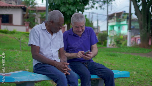 Two diverse seniors looking at phone sitting on park bench outdoors laughing and smiling together. Older friend sharing online media content to companion depicting authentic friendship © Marco