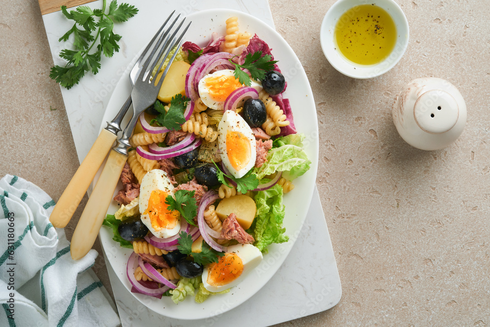 Tuna salad with pasta, eggs, potatoes, olives, red onions and sauce in white plate on old light gray concrete table background. Nicoise salad. French cuisine. Top view. Flat lay. Tuna salad with pasta, eggs, potatoes, olives, red onions and sauce in white plate on old light gray concrete table background. Nicoise salad. French cuisine. Top view. Flat lay.