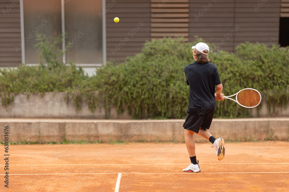 A teenage boy in a black t-shirt hitting the tennis ball on the court