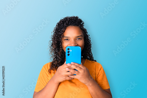 Young black woman holding her smartphone in front of her face to hide her smile with blue background in background. Smartphone camera.