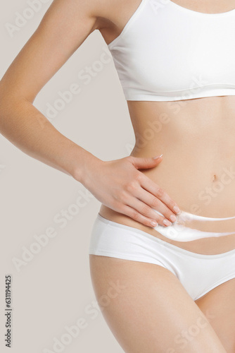 Young skinny girl in white lingerie on beige background applies cream on her belly