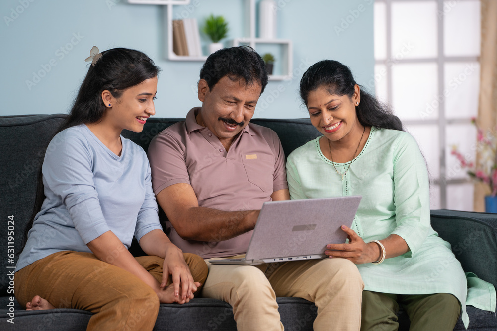Obraz premium Happy indian middle aged parents with adult daughter using laptop on sofa at home - concept of Modern Family, Togetherness and Parental Support