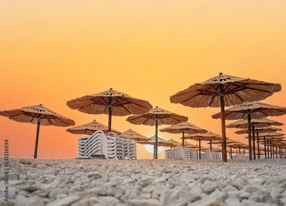 Straw umbrellas with sunbeds on the beach. Straw umbrellas and stacked deck chairs on the beach at sunset or sunrise.